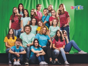 Zdjęcie zbiorowe Szczecińskiej Grupy Artystycznej Arfik. Widać na nim większą grupę członków zespołu, stojących i siedzących na zielonym tle. Wszyscy ubrani są w różnokolorowe podkoszulki z nazwą zespołu - napisem Arfik.
