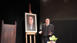 Zdjęcie z próby do spektaklu "Burmistrz". Widać na nim mężczyznę, aktora - Adama Dzieciniaka, stojącego obok swojego portretu. Obraz jest duży i stoi na sztaludze. Obok stoi też duże, ciemno brązowe, drewniane krzesło. Widać też kawałek kwiatka