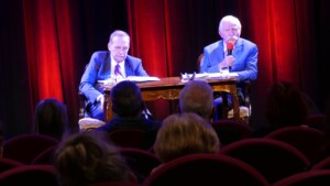 Zdjęcie z Zamkowych Spotkań z Psychologią i Sztuką. Na fotografii dwóch mężczyzn siedzących na scenie przed publicznością, jeden z nich trzyma mikrofon.