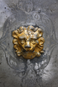 Fragment sarkofagu książęcego. Na szarym tle rzeźbiona złota głowa lwa.