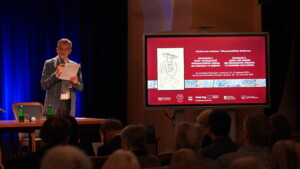 Moderator konferencji prof. Paweł Gut. Obok niego ekran, na którym wyświetlany jest plakat konferencji.