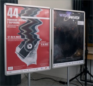 Zdjęcie dwóch stojaków z plakatami. Jeden w kolorze czerwonym promuje 44. Pomorskie Spotkania z Diaporamą. Drugi w ciemnym kolorze reklamuje wystawę WieloŚwiaty.